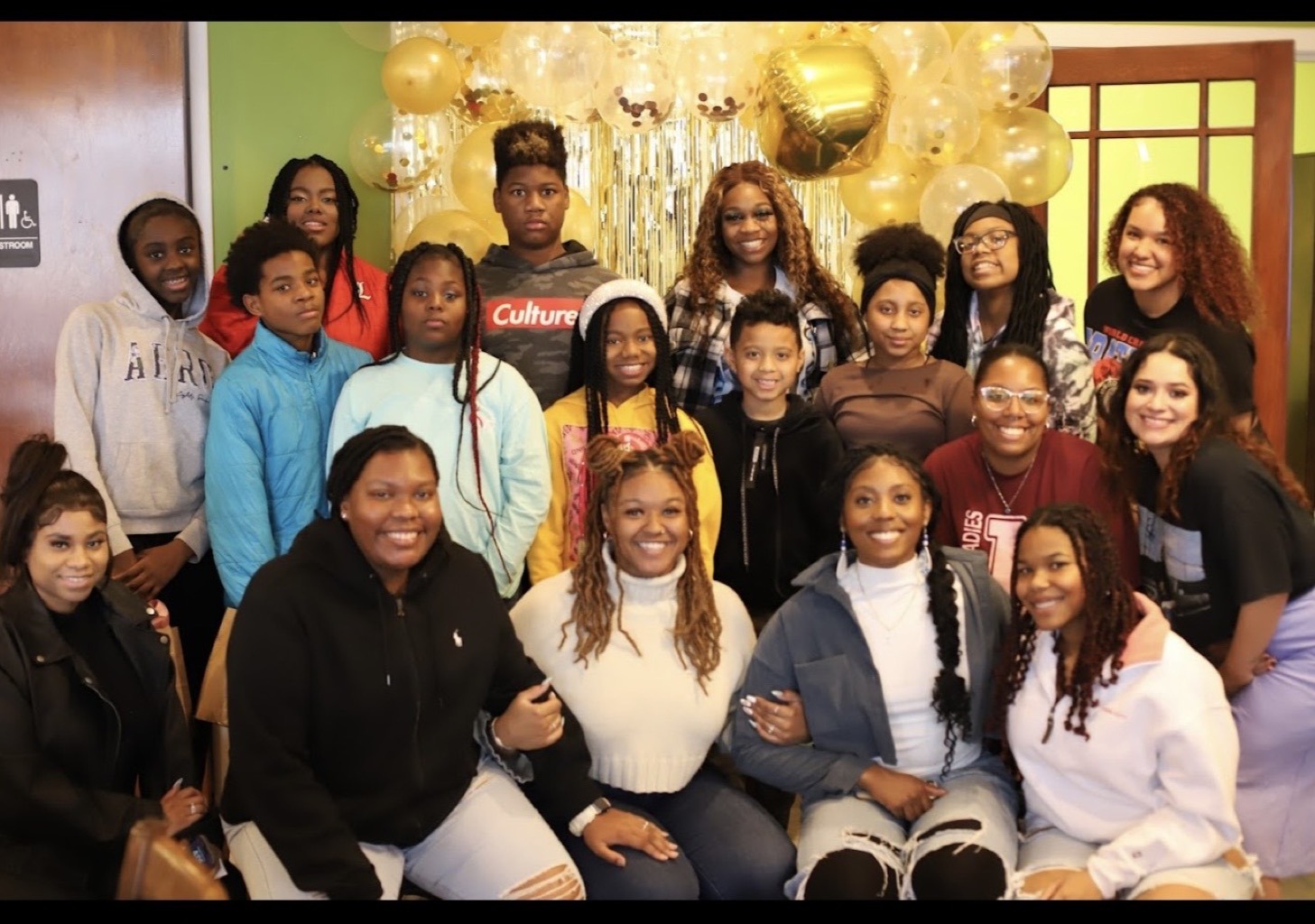 A large group of teens and young adult mentors posing together at an indoor Huddle celebration gathering with gold balloons