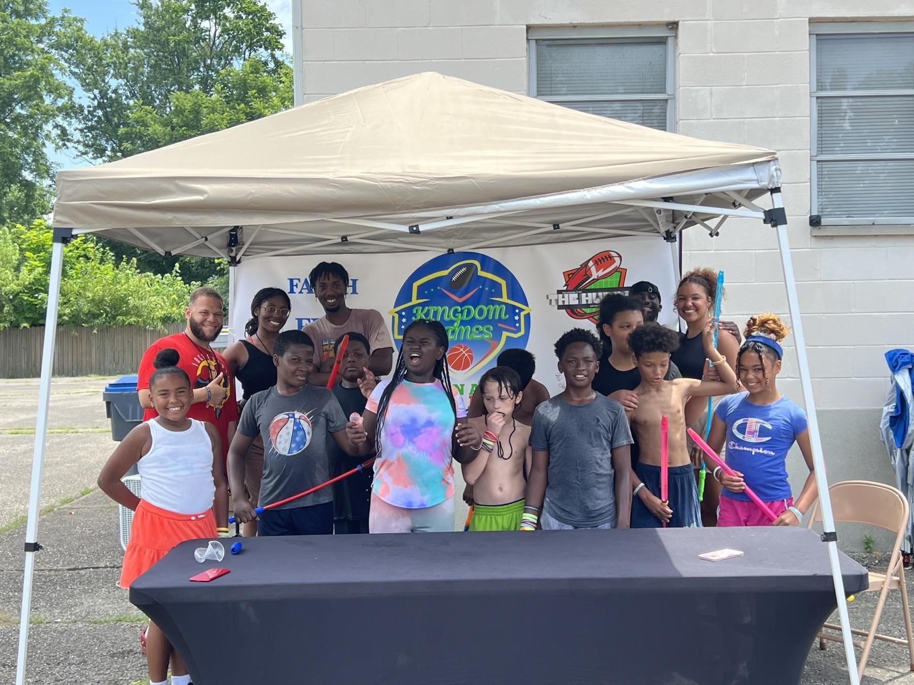 Kids and Huddle volunteers gathered under a tent in front of the Kingdom Games Faith in Action Field Day banner and The Huddle logo