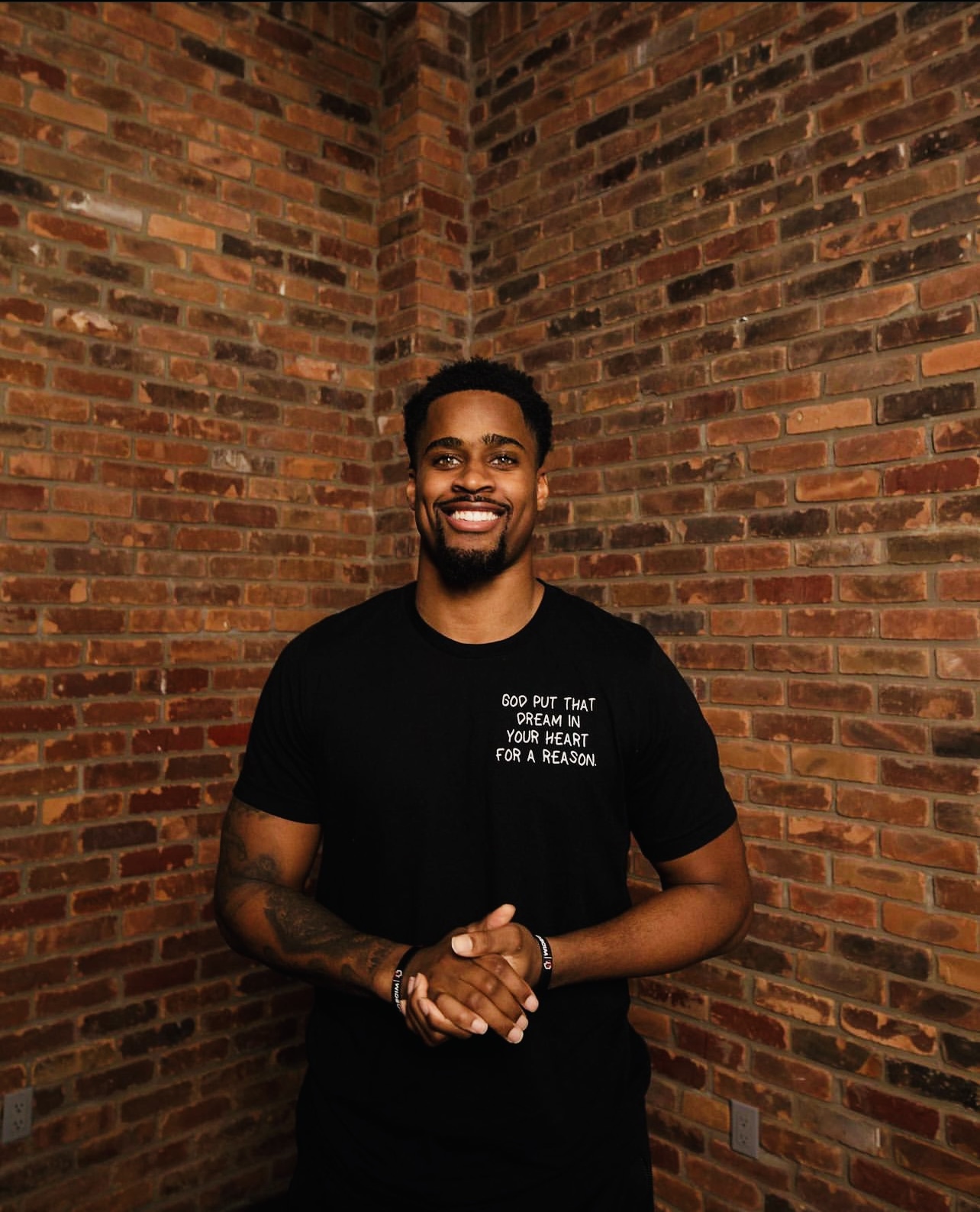Daylen Baldwin, Executive Director of The Huddle Inc., smiling in front of a brick wall wearing a black t-shirt