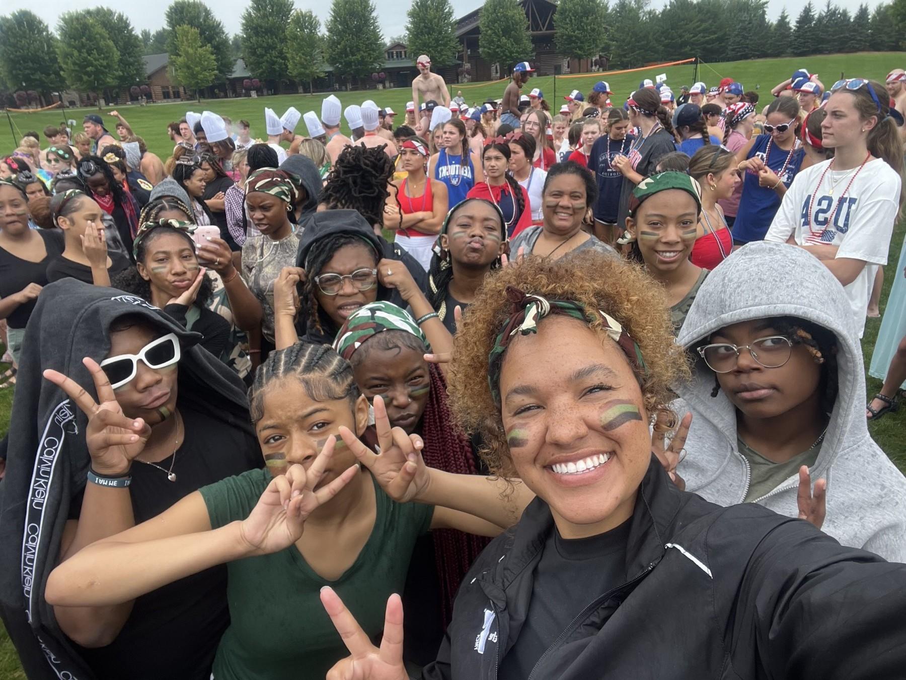 A camp field-day group selfie of Huddle campers and staff wearing face paint and camo bandanas on a grass field
