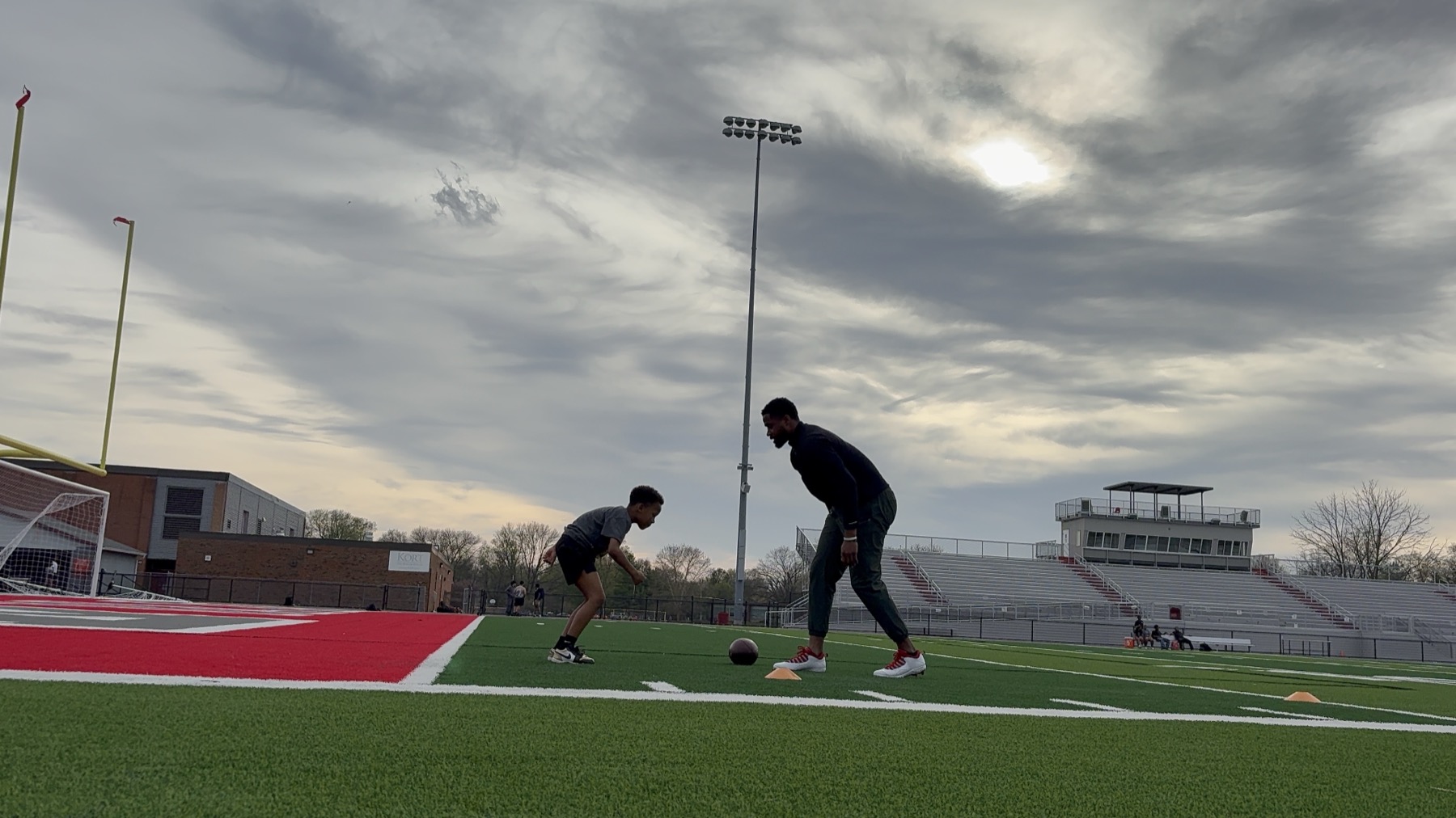 A Huddle coach training a young athlete on an outdoor football field under an overcast sky